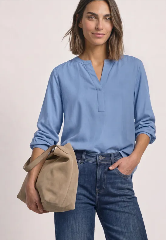 Frau in hellblauer Bluse und Jeans hält eine beige Tasche, vor schlichter Kulisse.