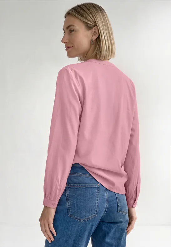 Eine Frau im Profil trägt eine rosa Bluse mit langen Ärmeln und blaue Jeans vor hellem Hintergrund.