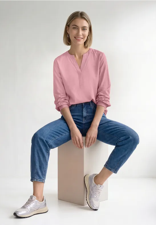 Eine Frau sitzt lächelnd auf einem Würfel, trägt pinke Bluse, blaue Jeans und weiße Sneaker vor weißem Hintergrund.
