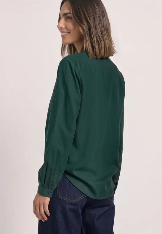 Frau in dunkelgrüner Bluse und Jeans, seitlich stehend und lächelnd.