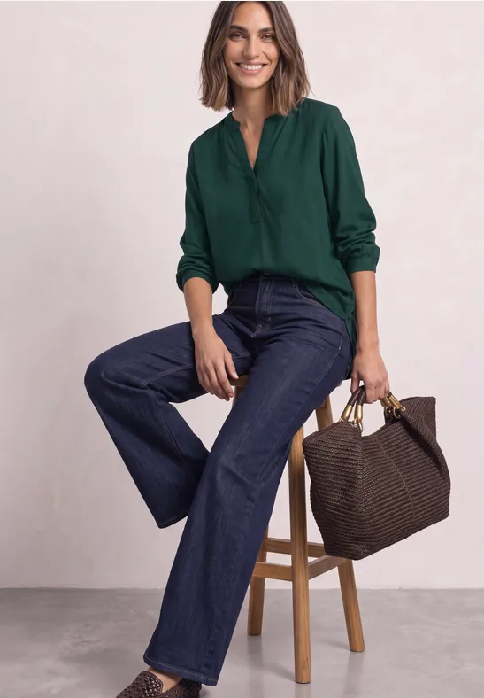 Lächelnde Frau mit grünem Blusenoberteil und weiten Jeans sitzt auf Hocker und hält braune Tasche.