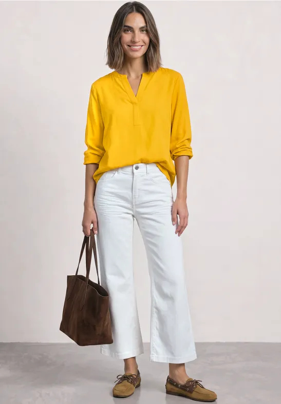 Frau in gelber Bluse, weißen Culotte-Jeans, braunen Loafers und Wildledertasche steht vor hellem Hintergrund.