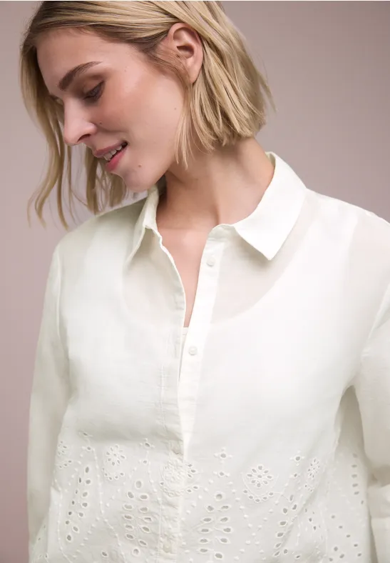 Vrouw draagt witte blouse met opengewerkt borduurwerk, kraag en knoopsluiting, deels transparant.