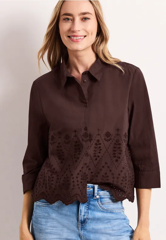 Femme en blouse marron foncé à broderies ajourées, ourlet festonné et manches 3/4, portée avec un jean.