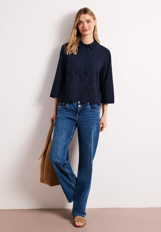 Vrouw in marineblauwe kanten top met driekwartmouwen, rechte jeans, sandalen en een bruine tas.