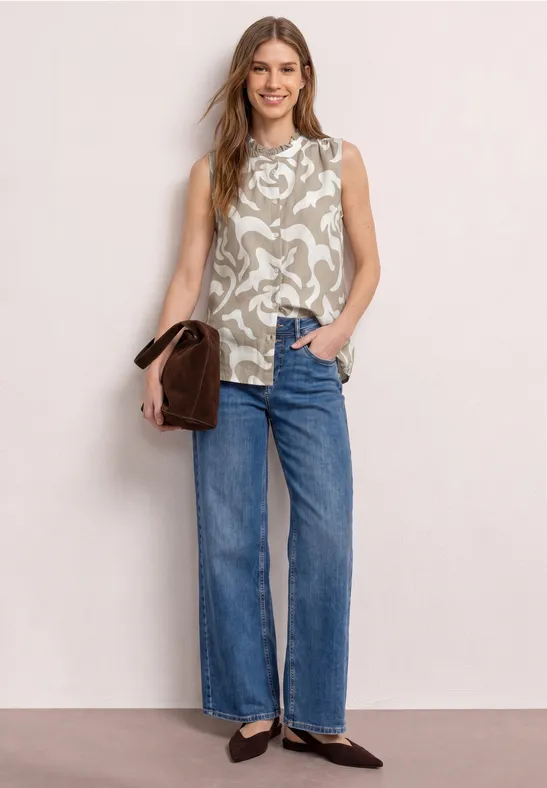 Vrouw in mouwloos dessin/top, rechte jeans, zwarte ballerina’s en bruine handtas.