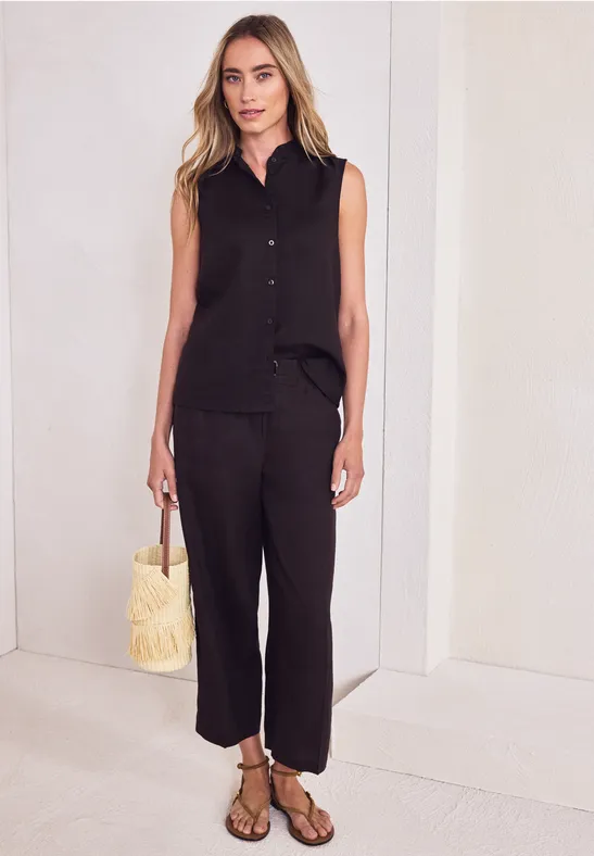 Frau in schwarzem ärmellosen Hemd, lockeren Hosen, Sandalen und Strohtasche vor weißem Hintergrund.