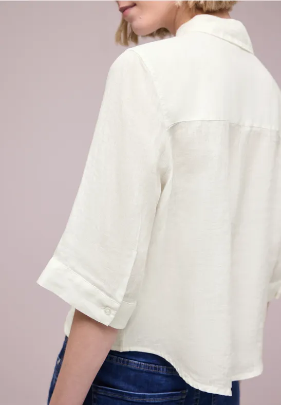 Frau im hellen, lockeren Hemd mit hochgekrempelten ¾-Ärmeln, Rückenansicht, trägt blaue Jeans.