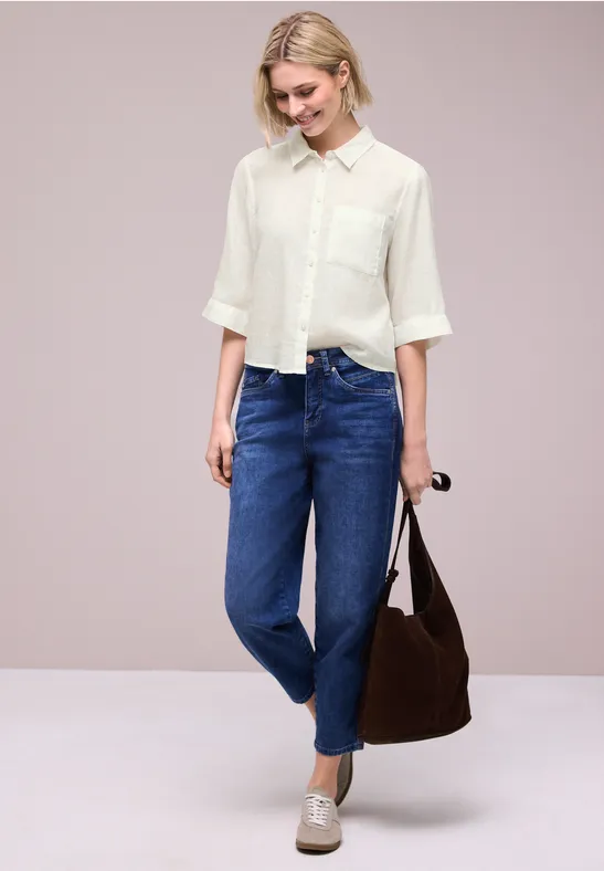 Frau in weißer Bluse und blauen Jeans steht mit brauner Tasche und Sonnenbrille in der Hand vor neutralem Hintergrund.