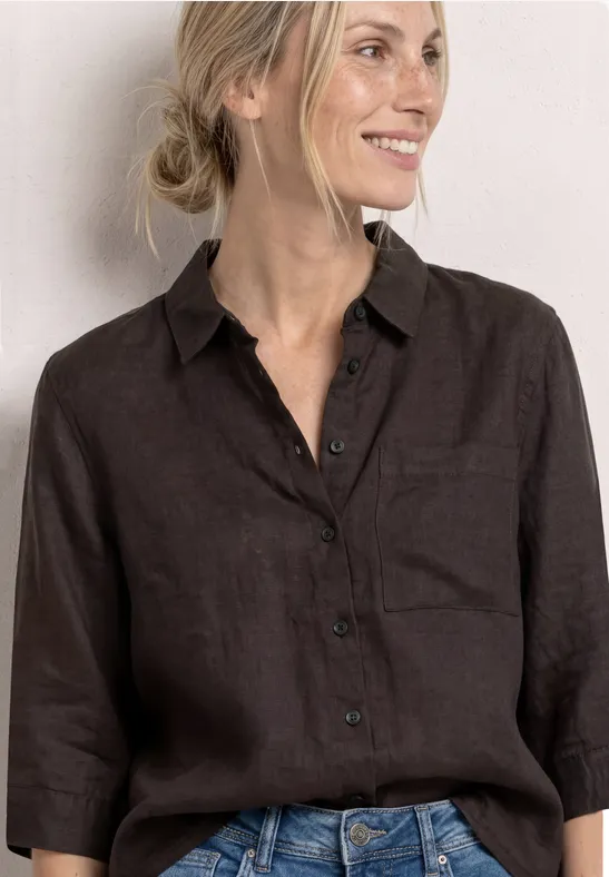 Frau in dunkelbraunem Leinenhemd mit 3/4-Ärmeln und Jeans vor hellem Hintergrund.