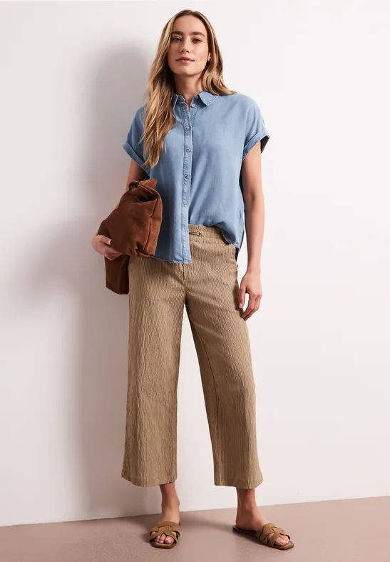 Vrouw in een casual zomerse outfit met lichtblauw bloesje, beige culotte, bruine sandalen en bijpassende tas.