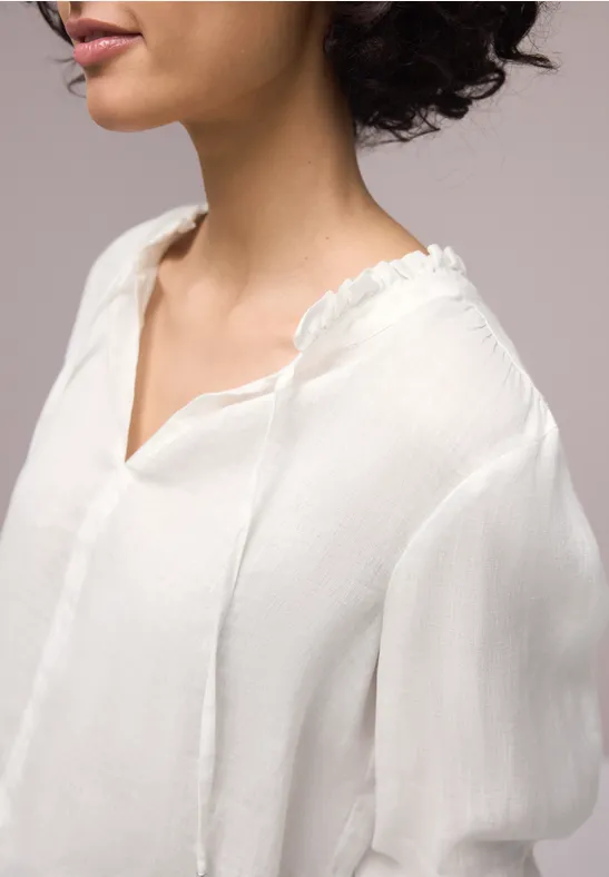 Een vrouw in profiel draagt een witte, lichte blouse met V-hals en losse, wijdvallende mouwen.