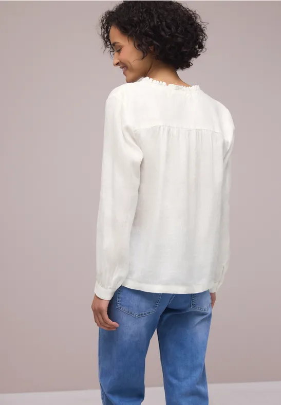 Vrouw van achteren in een wit bloesje en blauwe spijkerbroek tegen neutrale achtergrond.