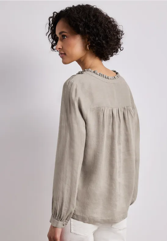 Vrouw van achteren in een losvallende grijze linnen blouse met plooien bij schouders en manchetten, lichte broek.