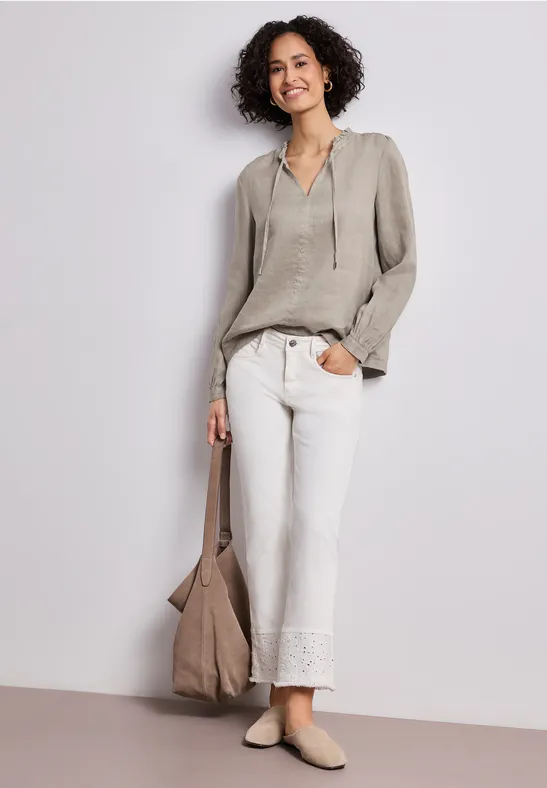 Persoon draagt lichtgrijs shirt, witte broek met kanten zoom, beige tas en platte schoenen tegen neutrale achtergrond.