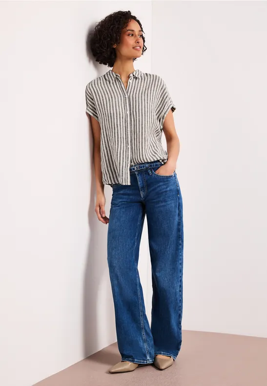 Une personne appuyée contre un mur, vêtue d’une chemise rayée et d’un jean bleu large.