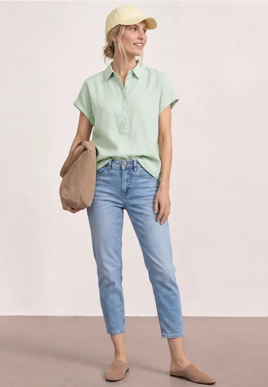 Vrouw in casual zomerse outfit: lichtgroene blouse, blauwe jeans, beige loafers en een tas.