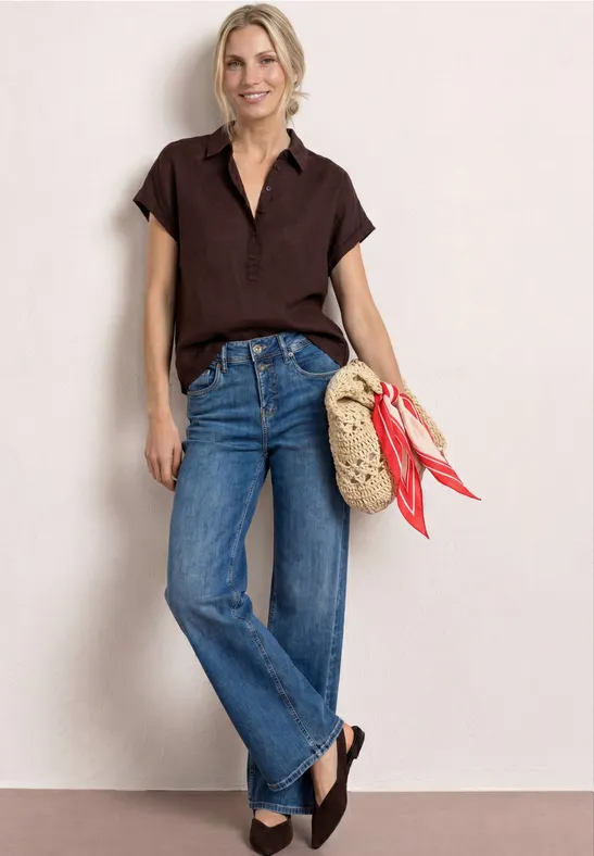 Femme en chemise marron courte et jean bleu, portant un panier en osier avec foulards rouges et des chaussures compensées.