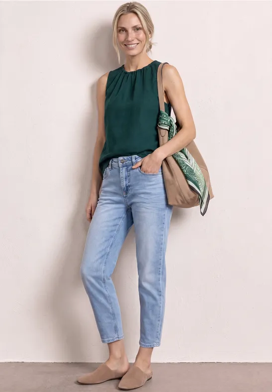 Eine Frau trägt ein dunkelgrünes, ärmelloses Top, helle Jeans, beigen Slipper und eine große Tasche.