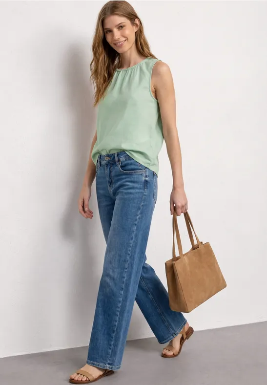 Frau trägt mintgrünes Top, weite Jeans, braune Sandalen und Handtasche vor weißer Wand.