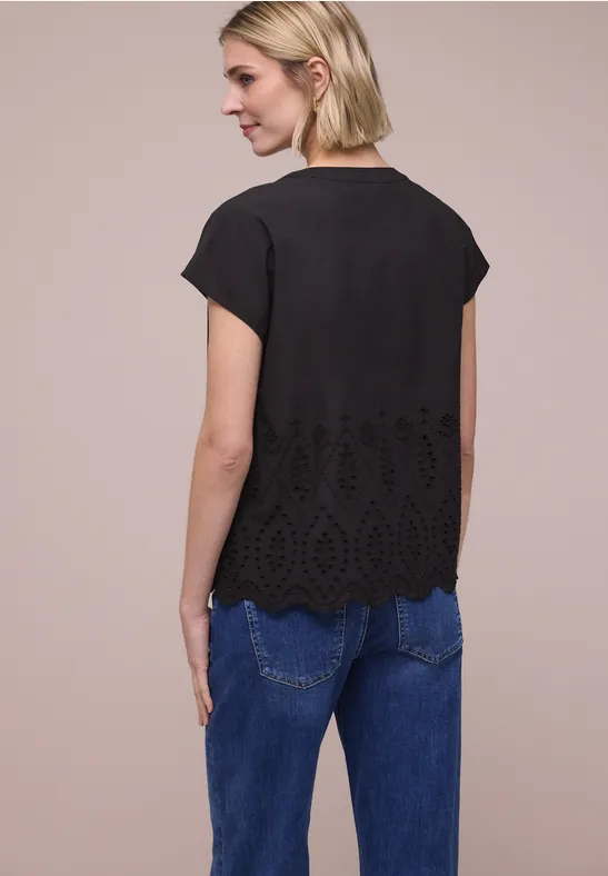 Vrouw van achteren in zwarte korte mouwenblouse met borduurwerk aan de zoom en blauwe spijkerbroek voor een beige achtergrond.