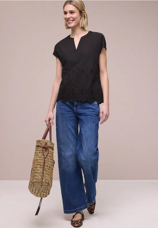 Vrouw met zwarte kanten top, wijdvallende jeans, luipaardballerinas en gevlochten tas tegen een beige achtergrond.