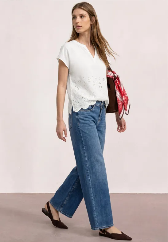 Vrouw in witte korte mouwen blouse en wijde blauwe jeans, zwarte platte schoenen, met een rood sjaaltje.