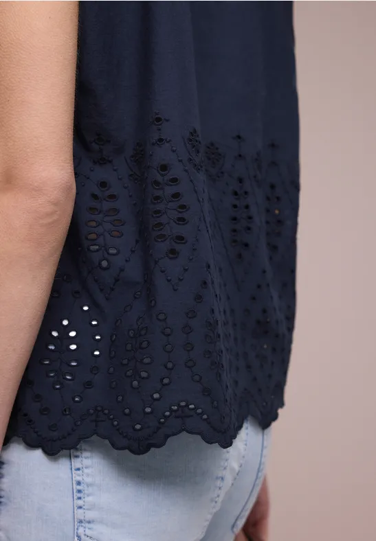 Persoon in lichte spijkerbroek en donkerblauw mouwloos topje met opengewerkt borduurwerk en gegolfde zoom.