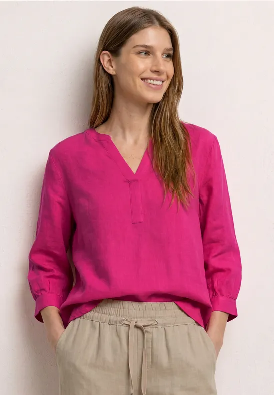 Vrouw in roze bloes en beige broek, ontspannen pose tegen lichte achtergrond.