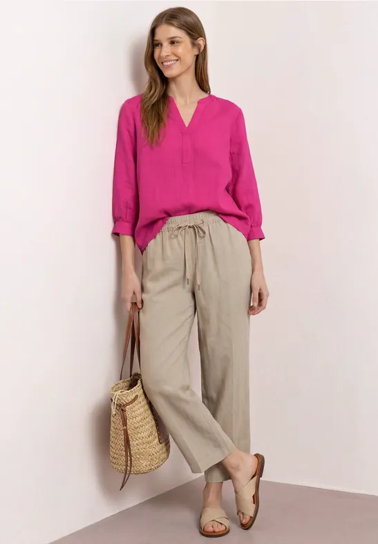 Een vrouw draagt een roze blouse, beige linnen broek, sandalen en een gevlochten strooien tas.