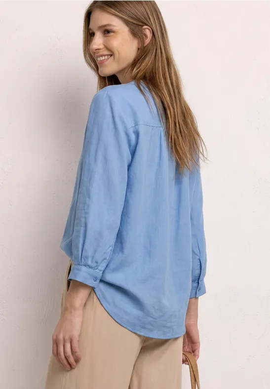 Zijaanzicht van een vrouw in een lichtblauw overhemd, beige broek en met een klein tasje.