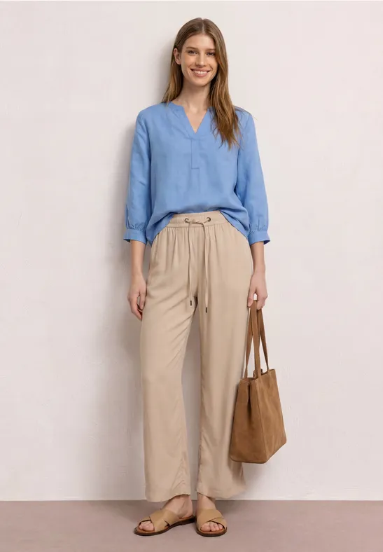 Vrouw met lichtblauwe blouse, beige paperbag-broek, platte sandalen en een bruine suède tas.