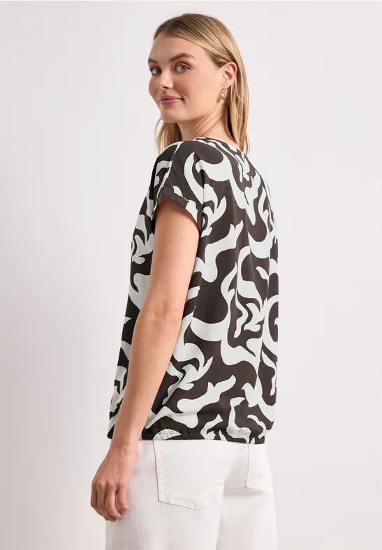 Vrouw in profiel met zwart-wit patroonshirt met korte mouwen en witte broek tegen een neutrale achtergrond.