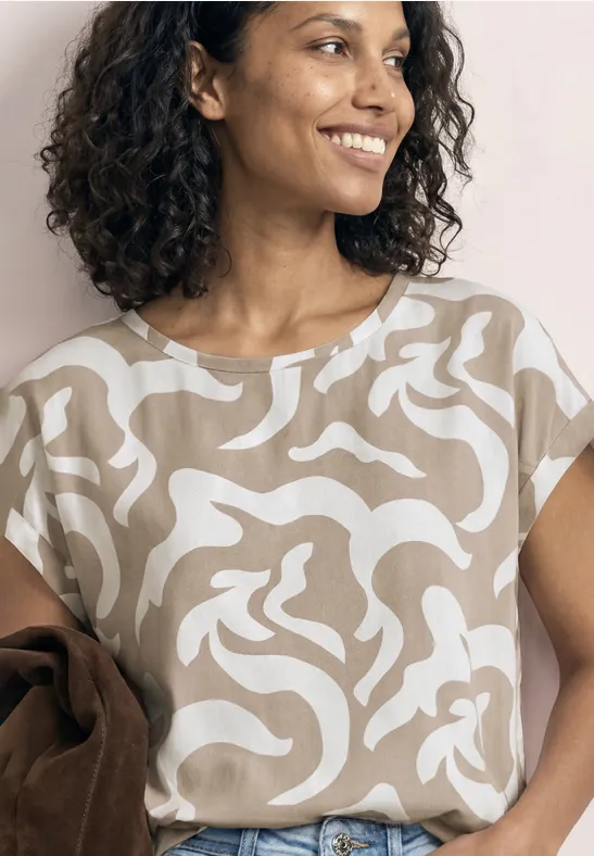 Eine Frau mit lockigem Haar trägt ein beigeweiß gemustertes Shirt und hält eine braune Jacke über dem Arm.