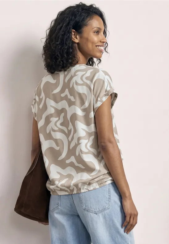 Eine Frau von hinten trägt ein beige-weiß gemustertes Shirt, hellblaue Jeans und eine braune Tasche.