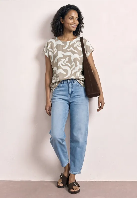 Eine Person trägt ein gemustertes T-Shirt, hellblaue Jeans mit Umschlag und schwarze Sandalen, dazu eine braune Tasche.