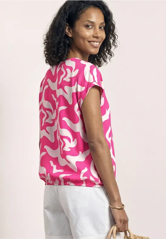 Frau von hinten mit pink-weiß gemustertem Top, weißer Hose und Basttasche.