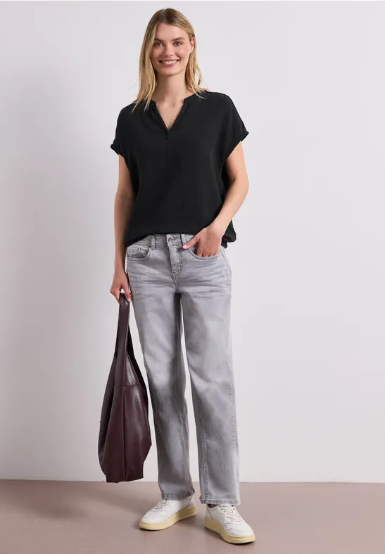 Vrouw in zwarte V-hals top, grijze wijde jeans, witte sneakers, met een grote bordeauxrode tas.