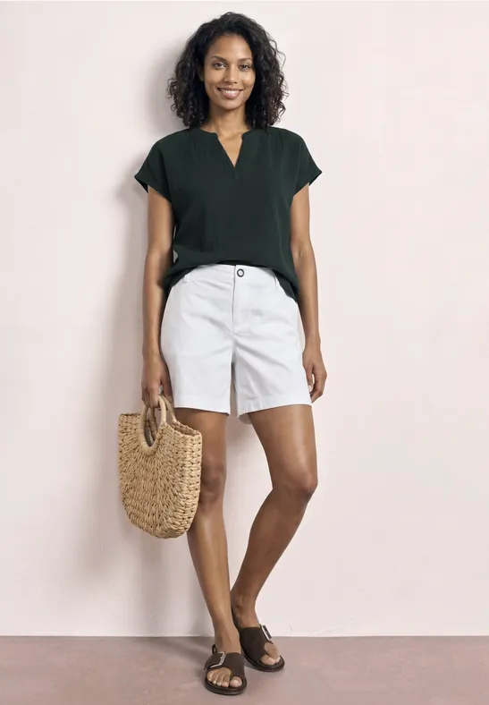 Model draagt zwart T-shirt, witte short, zwarte sandalen en een strooien tas tegen lichte achtergrond.