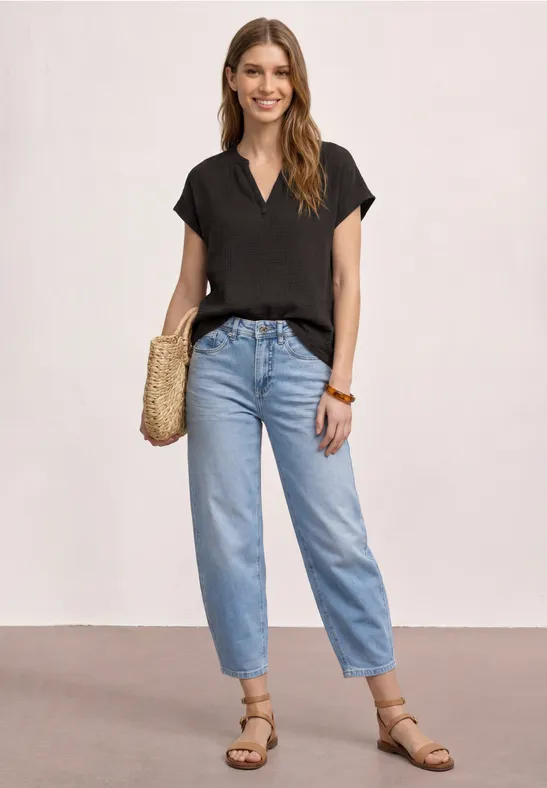 Femme en t-shirt noir, jean mom bleu clair, sandales plates et sac en paille sur fond uni.