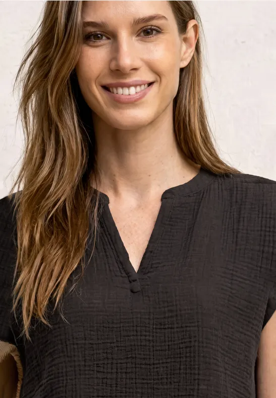 Femme aux cheveux bruns longs porte un top noir texturé sans col avec encolure fendue.