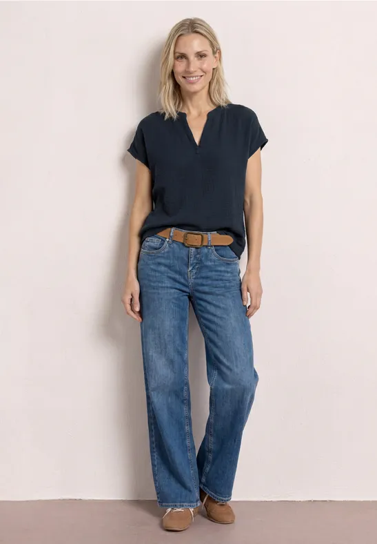 Frau vor heller Wand mit dunkelblauer Bluse, geraden Jeans, braunem Gürtel und flachen Schuhen.