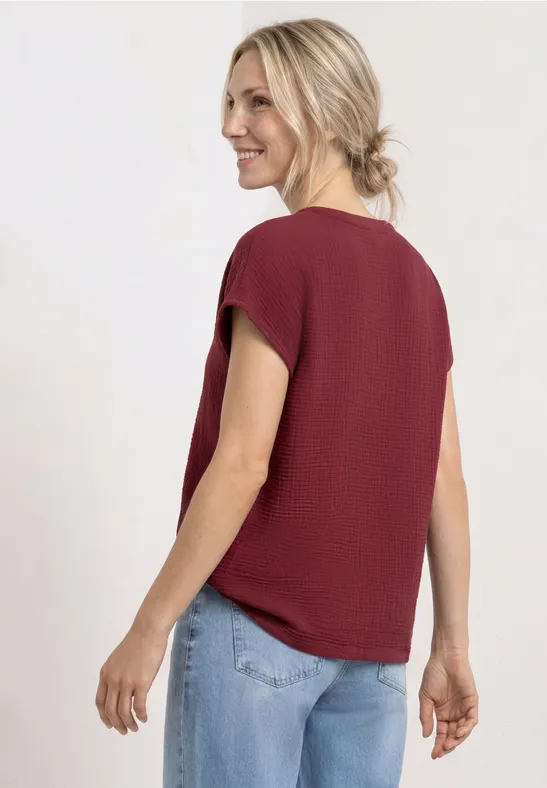 Frau von hinten in locker sitzendem bordeauxrotem Shirt und hellblauen Jeans vor neutralem Hintergrund.