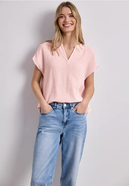 Vrouw in een roze top en blauwe jeans staat nonchalant met haar handen in de zakken voor een neutrale achtergrond.