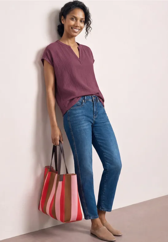 Femme en haut violet, jean bleu et sac rayé coloré devant un fond clair.