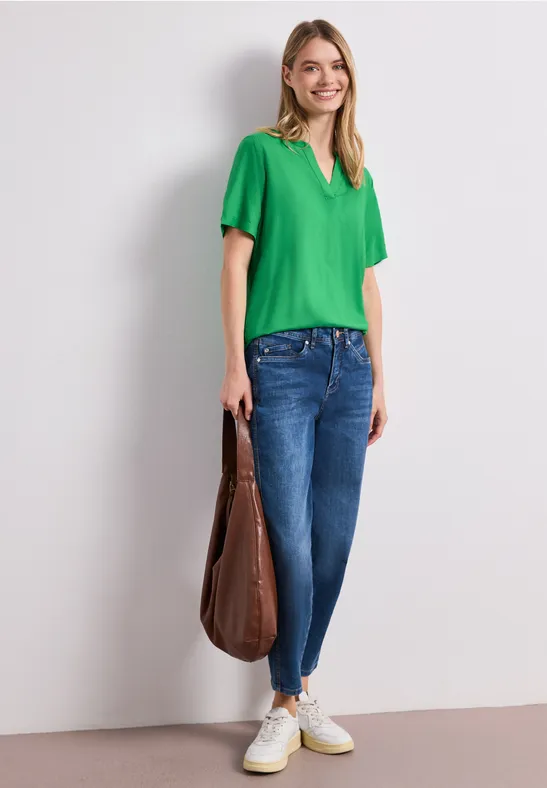 Frau trägt grünes T-Shirt, blaue Jeans, weiße Sneaker und große braune Tasche vor hellem Hintergrund.