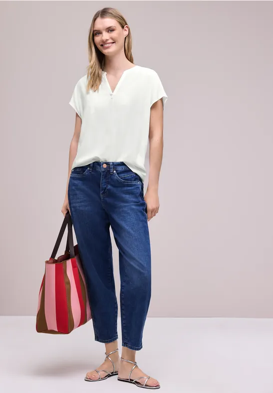 Frau trägt weiße V-Ausschnitt-Bluse, blaue Mom-Jeans, silberne Sandalen und gestreifte Tragetasche.