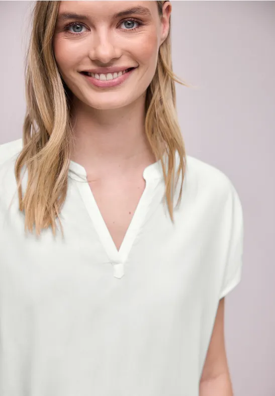 Eine Person trägt eine schlichte weiße Bluse mit V-Ausschnitt vor neutralem Hintergrund.