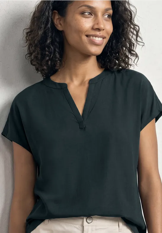 Eine lächelnde Frau trägt eine schwarze Kurzarmbluse mit V-Ausschnitt vor neutralem Hintergrund.