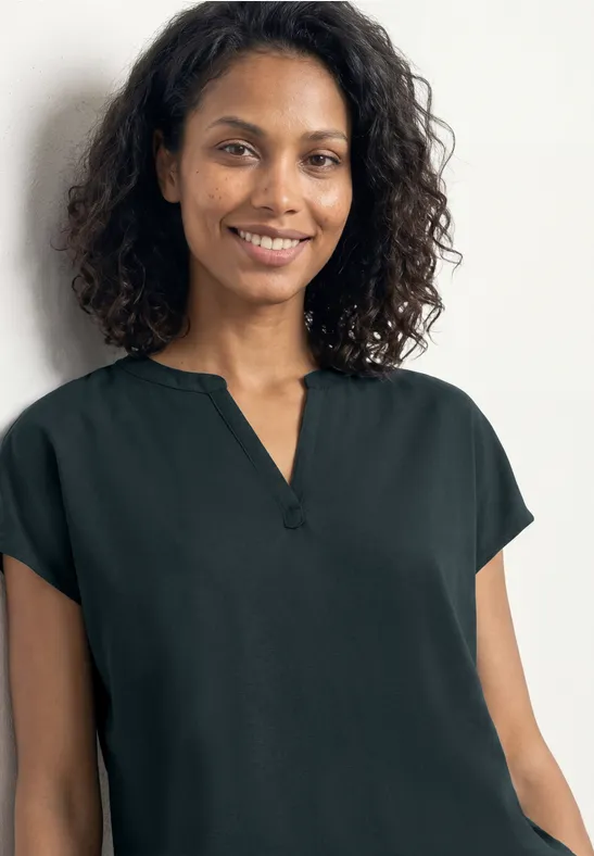 Frau mit lockigem Haar trägt schlichte, dunkelgrüne Kurzarmbluse vor weißem Hintergrund.